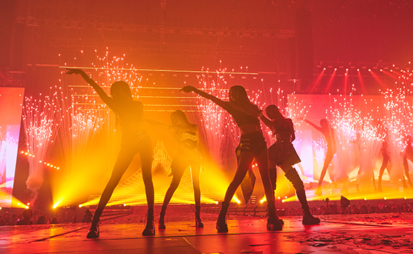 Silhouetted dancers perform on a stage illuminated by bright orange lights and falling sparks.