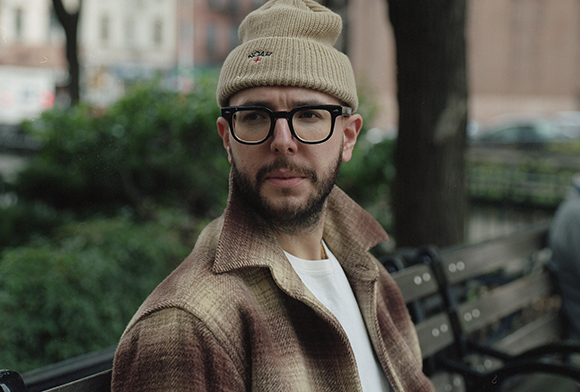 A man wearing black-rimmed glasses, a tan beanie, and a brown textured jacket sits on a park bench.