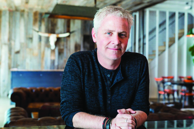 A man with short gray hair wearing a dark long-sleeved shirt leans on a table in a rustic interior.
