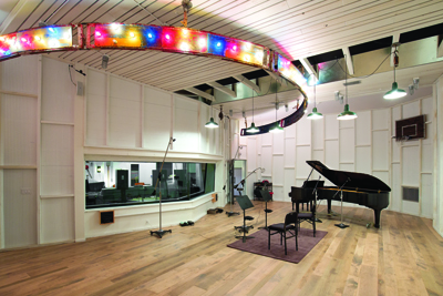 A large recording studio with light wood floors, white paneled walls, and a grand piano.
