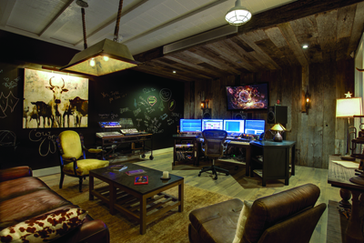 A rustic music studio with wooden walls, multiple computer monitors, a keyboard, and leather sofas.
