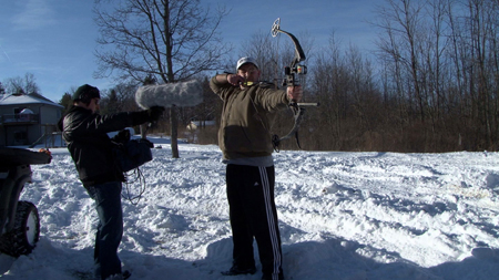 A man aims a compound bow in a snowy field while a person films him with a professional camera.