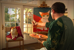 A man in a green shirt paints a portrait of an orange cat on an easel while looking at a real cat on a chair.