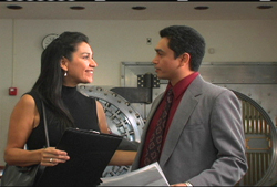 A woman in a black top and a man in a grey suit talk in front of a large metal vault door.