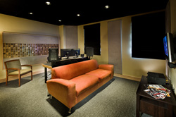An orange sofa sits in a room with a desk, computer monitors, and acoustic wall panels.