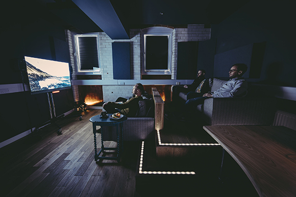 Three people sitting on couches in a dark room watching a large screen displaying a landscape.