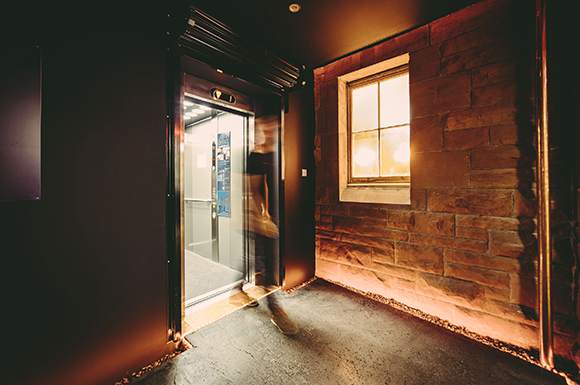 A blurred figure walks through an open elevator door next to a stone wall and a lit window.