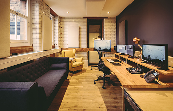 A dark sofa and yellow armchair face a long wooden desk with multiple computer monitors and office equipment.