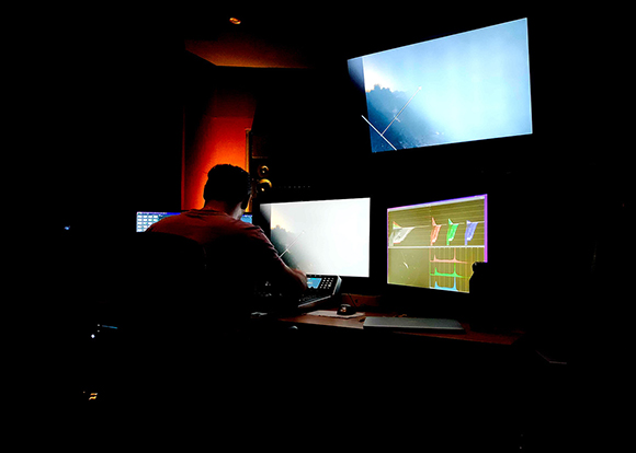 A person sits in a dark room facing three glowing computer monitors displaying various data and graphics.