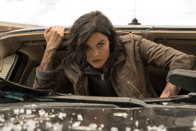A woman with dark hair and tattoos leans out from the open trunk of a dark car.