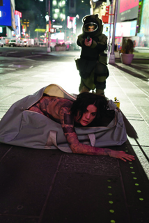 A tattooed person crawls from a large grey bag on a city sidewalk as a person in dark tactical gear aims a weapon.