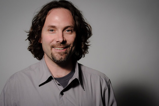 A man with wavy dark hair and a goatee smiling against a gray background.