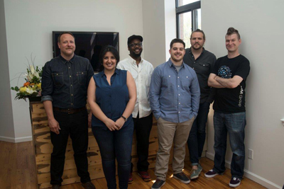 Six diverse adults standing in a row indoors against a white wall and a window.