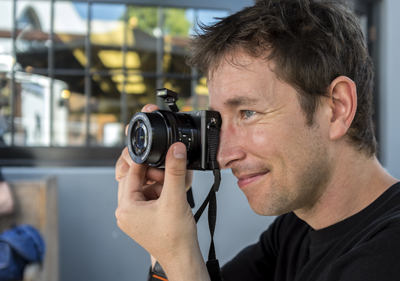 A man smiling while looking through the viewfinder of a black digital camera.
