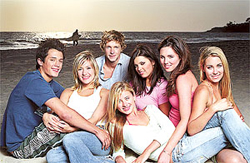 Seven young adults sit together on a sandy beach with the ocean and a surfer in the background.