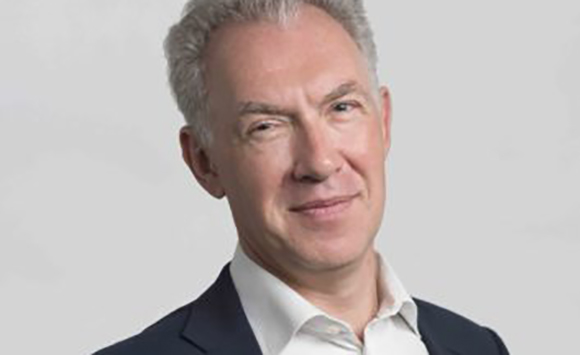 A middle-aged man with short grey hair smiling in a dark suit and white shirt against a plain background.