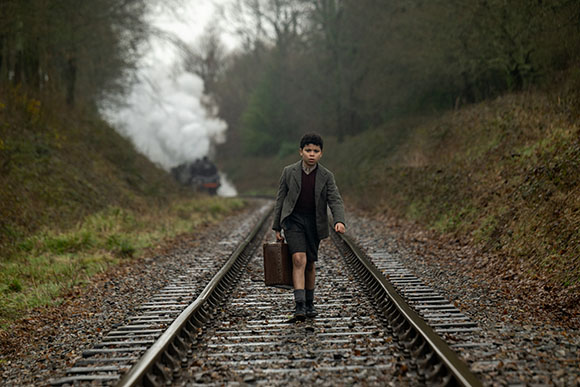 A young boy in a blazer and shorts walks along railroad tracks toward the camera with steam rising behind him.