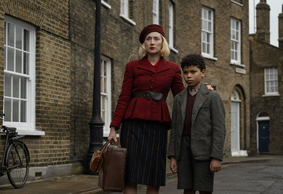 A blonde woman in a red jacket and beret stands with her hand on a young boy's shoulder in a brick street.
