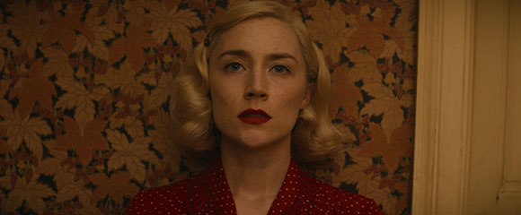 A blonde woman with red lipstick looks directly at the camera against patterned floral wallpaper.