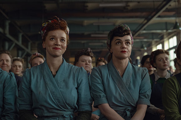 Two women in blue wrap-style work uniforms and headscarves look upward in a large industrial room.
