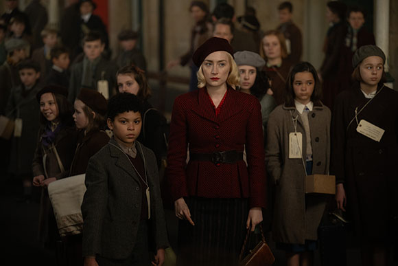 A woman in a red jacket and beret stands among a group of children in period clothing.