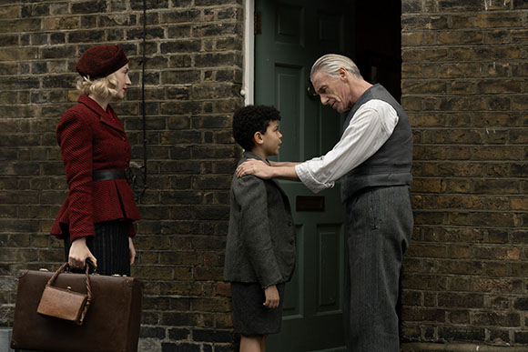A woman in a red jacket and a man in a grey vest stand by a doorway with a young boy.