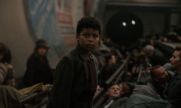 A young boy in a dark jacket looks back while standing in a crowded, dimly lit subway station.