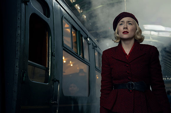 A blonde woman in a red beret and striped coat looks upward beside a dark train car.