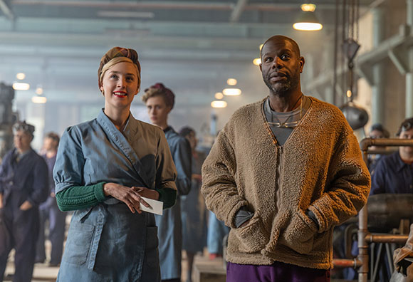 A woman in a blue uniform and a man in a brown fleece sweater stand in a factory.