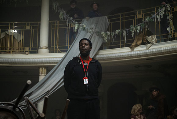 A man in a black hoodie stands in a dark, tiered room with white fabric draping from a balcony.
