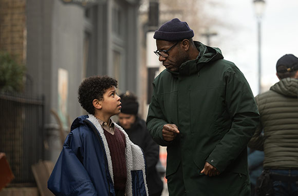A man in a green parka leans down to speak to a young boy in a blue jacket on a city street.
