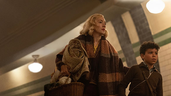A blonde woman in a patterned shawl and a young boy look upward in a dimly lit room.