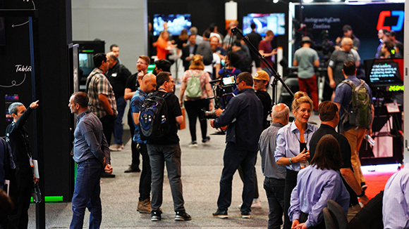 People at an exhibition hall, some looking at displays, others talking. A camera rig is visible.