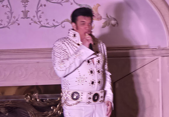 A man in a white, sequined jumpsuit and a wide belt, styled like Elvis Presley, sings into a microphone.