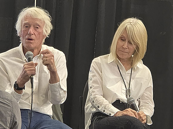 A man with white hair speaks into a microphone while a woman with blonde hair looks down, both wearing white shirts.