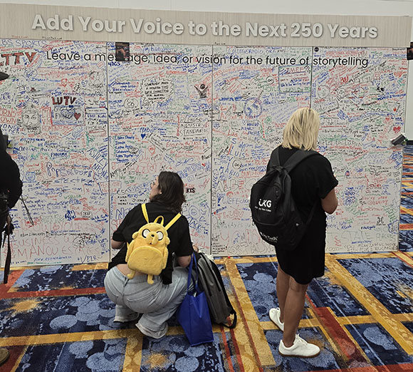 Two people observe a large white wall covered in colorful handwritten messages and drawings, with the text "Add Your Voice to