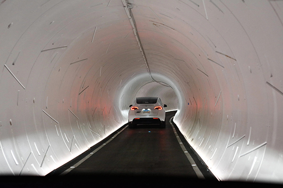 A white electric car drives down a tunnel with white walls and illuminated strips along the sides. The tunnel curves ahead.