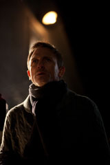 A man wearing a dark coat and scarf looks upward into a beam of light against a dark background.