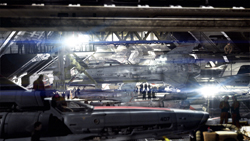 Several fighter jets parked inside a large, dimly lit aircraft hangar with bright overhead lights.