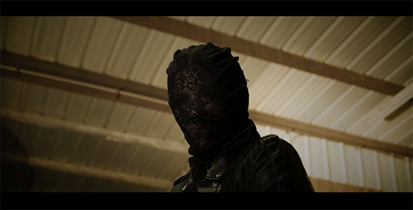 A person in a black lace mask and dark clothing looking downward against a corrugated ceiling.