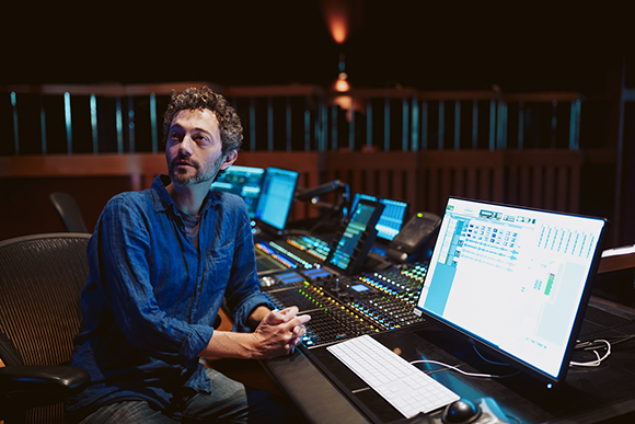 A man with curly hair sits at a large audio mixing desk with multiple monitors in a dark studio.