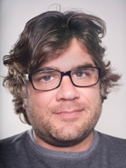A man with wavy brown hair and black-rimmed glasses looking forward.