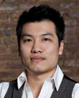 A man with short black hair wearing a white collared shirt and a dark grey vest against a brick wall.