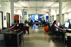 People working at desks in a bright, open-plan office with high ceilings and a ping pong table in the center.