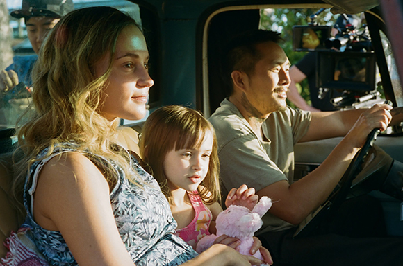 A woman and young girl sit in a car while a man drives, with film equipment visible in the background.