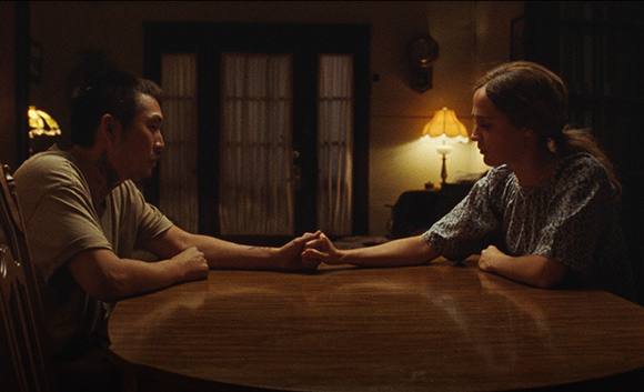A man and a woman sit across from each other at a wooden table, holding hands in a dark room.