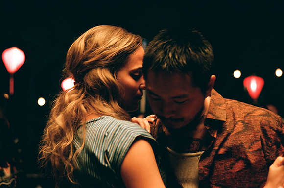 A man and a woman with long wavy hair lean toward each other in a dimly lit setting with red lanterns.