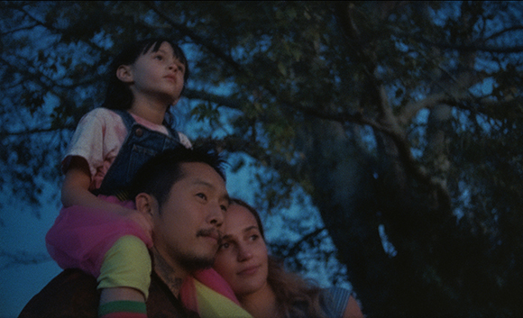 A man carries a young girl on his shoulders while a woman leans against him under dark trees.