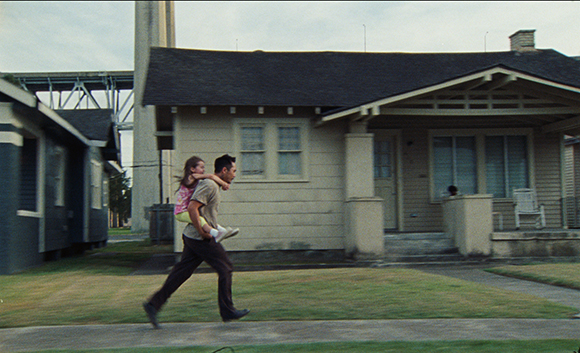 A man walks across a lawn carrying a young girl in a pink shirt and yellow shorts.