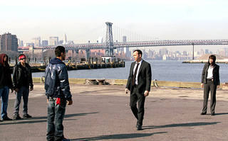 Five people stand on a waterfront pier with a large suspension bridge and city skyline behind them.